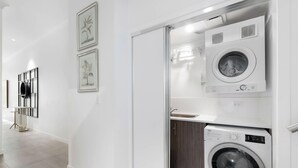 Ein moderner Waschraum mit zwei Waschmaschinen und einem Trockner, einem Metallgestell mit einem schwarzen Behälter sowie gerahmten Pflanzenbildern an der Wand.