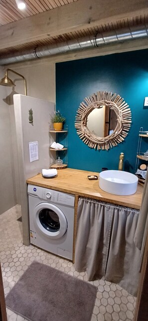 A modern bathroom with a washing machine, a round mirror, a sink, and a blue accent wall. - L'Oasis des Colibris – Tiny House & Spa in L'Étang-Salé (L'Étang-Salé)