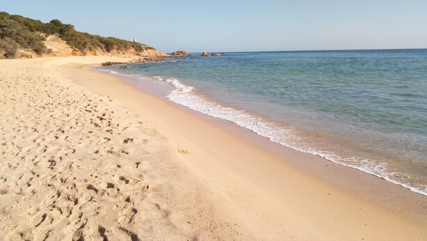 Una playa arenosa con huellas, cielo despejado y formaciones rocosas a lo lejos.
