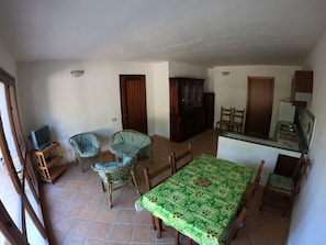 Un coin repas avec une nappe verte, des chaises en osier, un téléviseur posé sur un meuble en bois et un buffet.