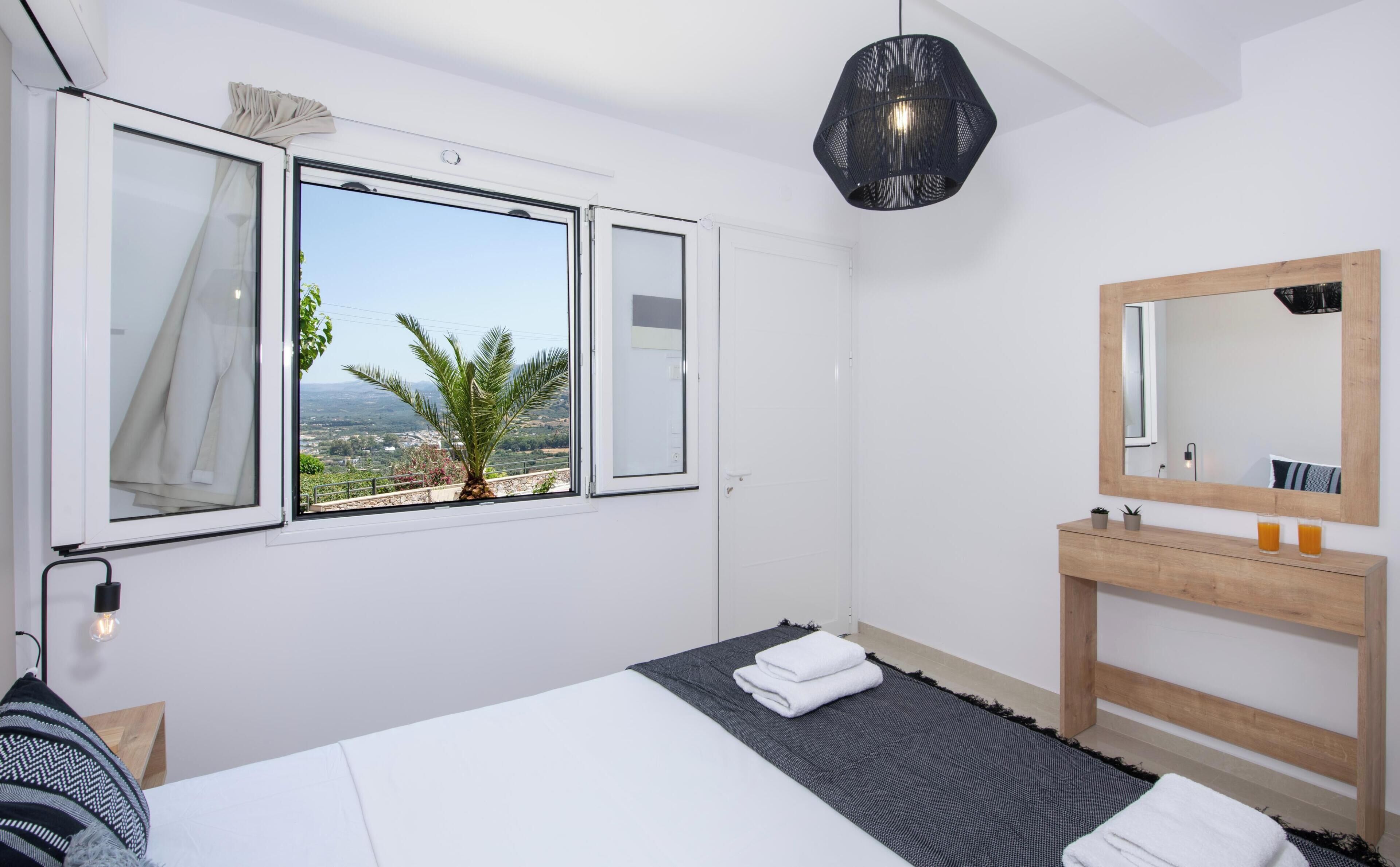 A bedroom with a bed, a window with curtains, a wooden bedside table with a mirror, and a view of a palm tree and landscape.