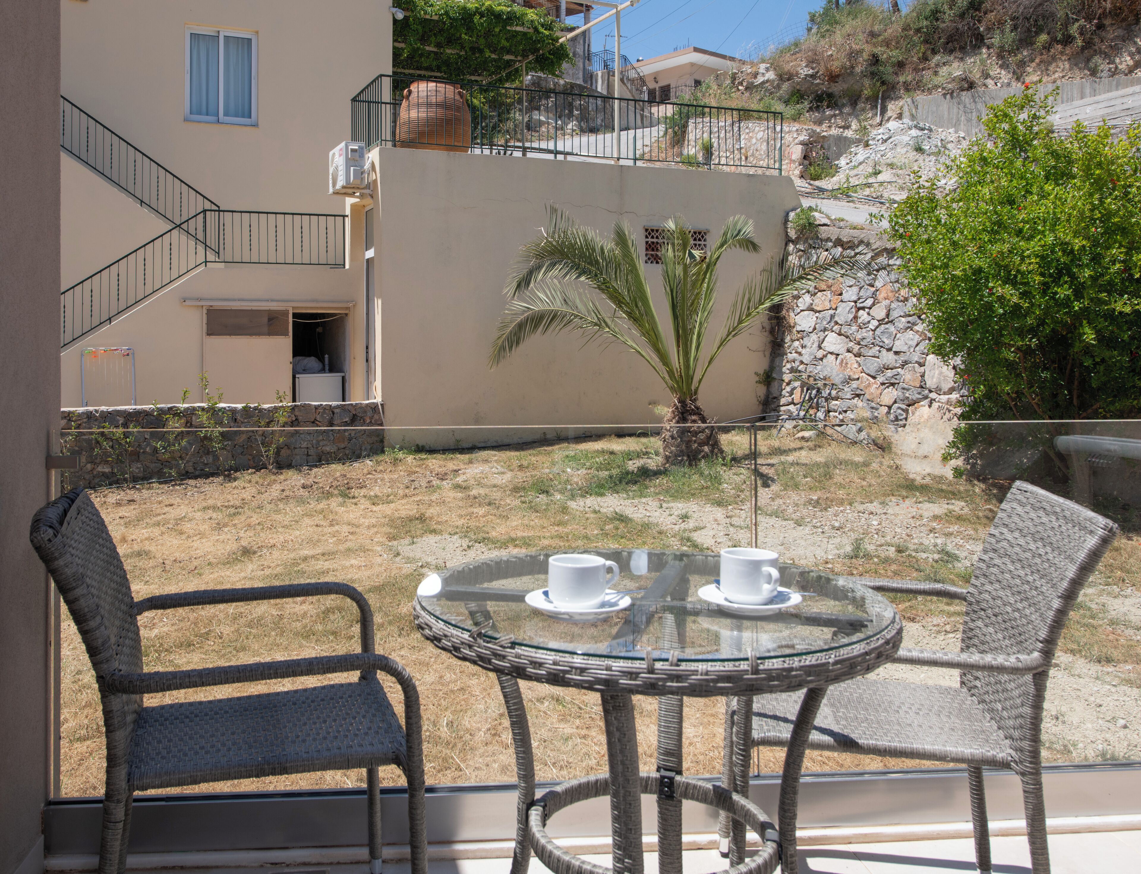 A balcony with wicker chairs and a glass table, a stone wall, and a view of a building with a staircase.