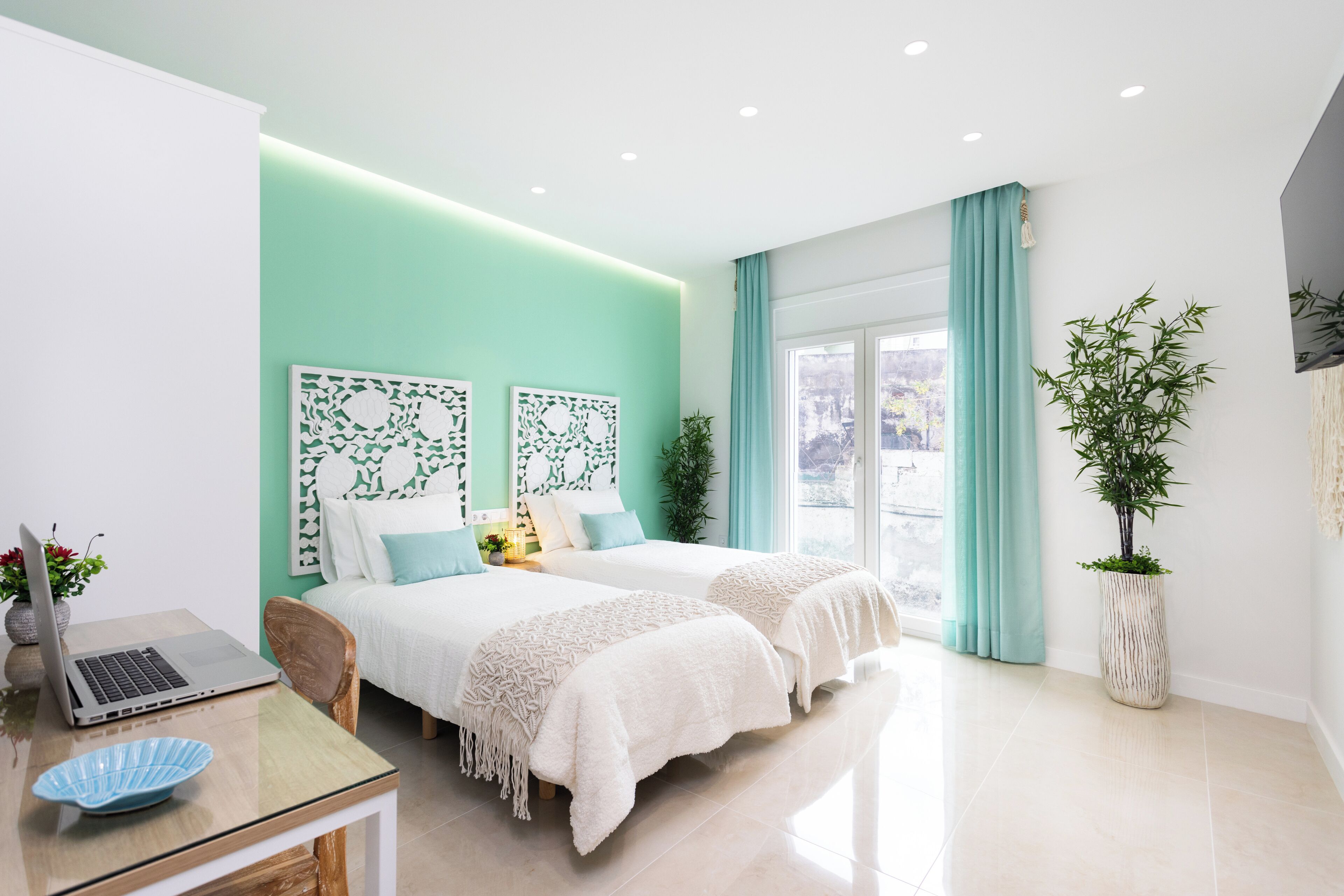 A modern bedroom with two beds, a desk with a laptop, and a large window with curtains.