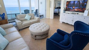 A spacious living room with a large window overlooking the sea, a sofa, a blue armchair, and a television on a white cabinet. - Sapphire - Just a Dream - 26th Floor Luxury (South Padre Island)