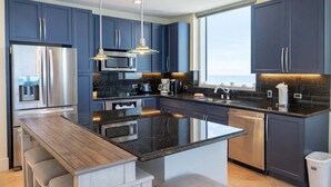 A modern kitchen with dark blue cabinets, stainless steel appliances, a large island with bar seating, and pendant lighting. - Sapphire - Just a Dream - 26th Floor Luxury (South Padre Island)