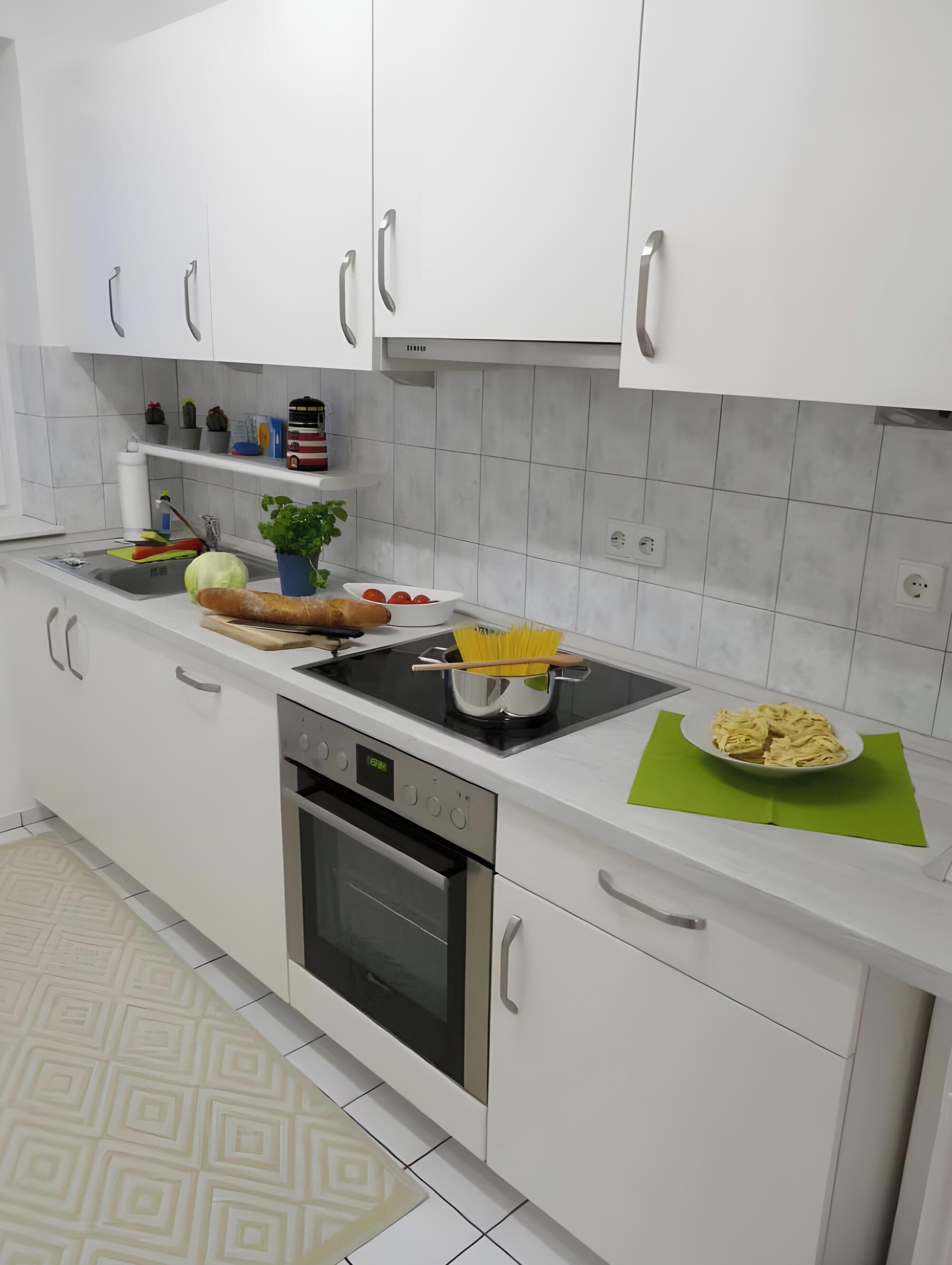 Et moderne køkken med hvide skabe, en indbygget ovn, en vask og diverse køkkenredskaber, som skærebræt, grøntsager og en skål med pasta.