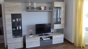 Una sala de estar moderna con un televisor de pantalla plana, armarios de madera y un estante pequeño con libros y objetos decorativos.