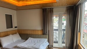 Une chambre d’hôtel avec un plafond en bois, un grand lit avec du linge de lit blanc, une vue sur la rue à travers une porte vitrée et un balcon avec des rideaux.
