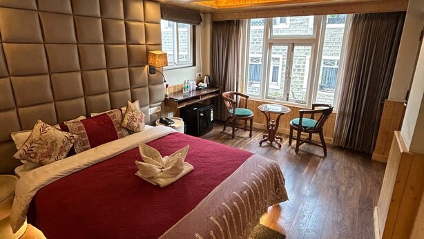 Une chambre d’hôtel avec un grand lit, un bureau et une chaise. Il y a des fenêtres avec des rideaux et une vue sur les bâtiments à l’extérieur.