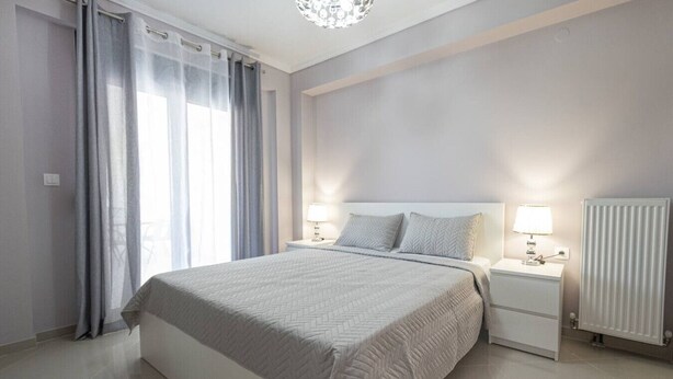 A modern bedroom with a large bed, two bedside tables with lamps, a radiator, and sheer curtains on a window. - Mpia House by RentalsPro (Nea Moudania)