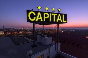 A neon sign reading "CAPITAL" with five stars above it, mounted on a rooftop with a view of a cityscape at dusk.