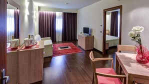 A hotel room with a wooden floor, a dining table with chairs, a TV, a desk with a computer, a sofa, and a red rug. - HOTEL CAPITAL (Rovigo)