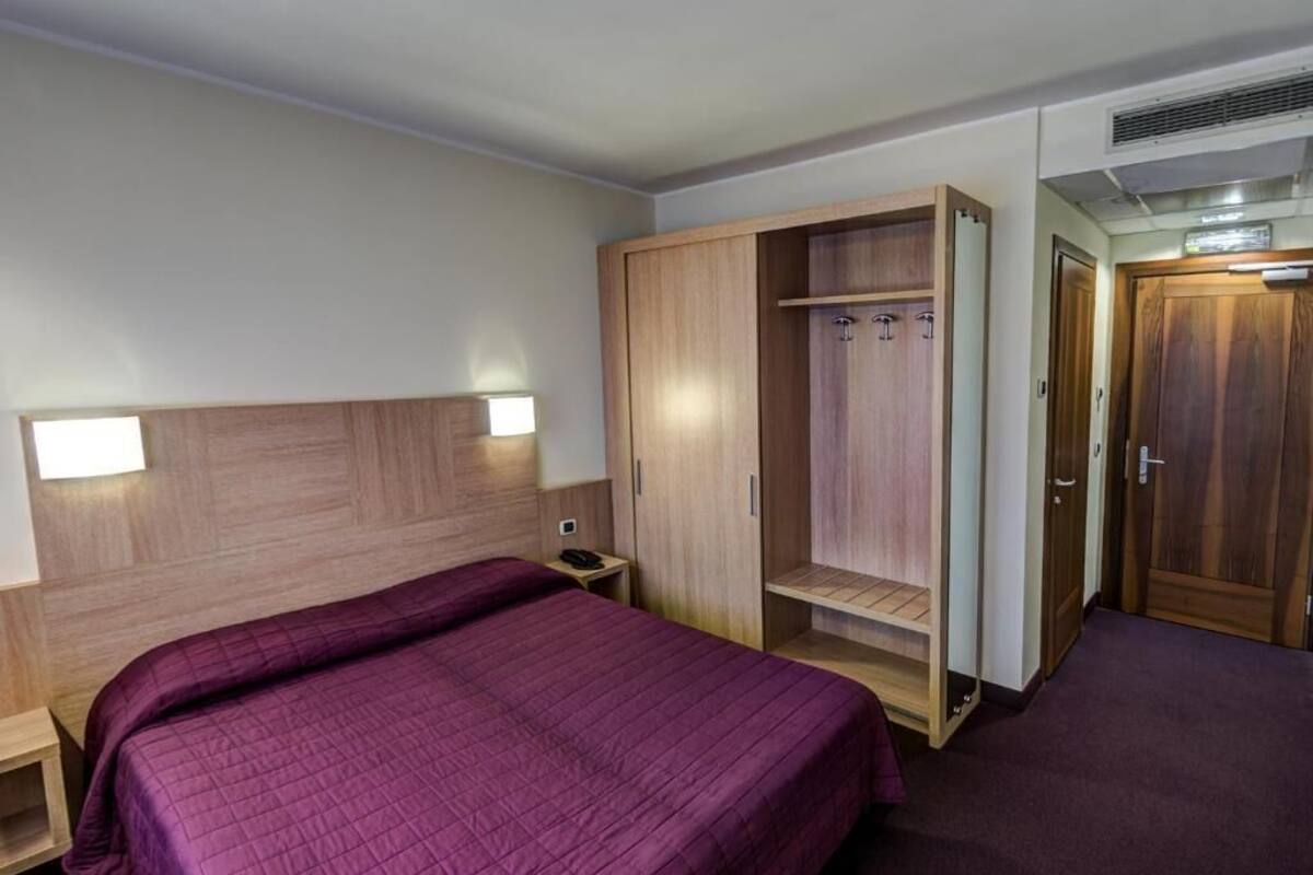 A hotel room with a bed, a wooden wardrobe, a nightstand, and a door.