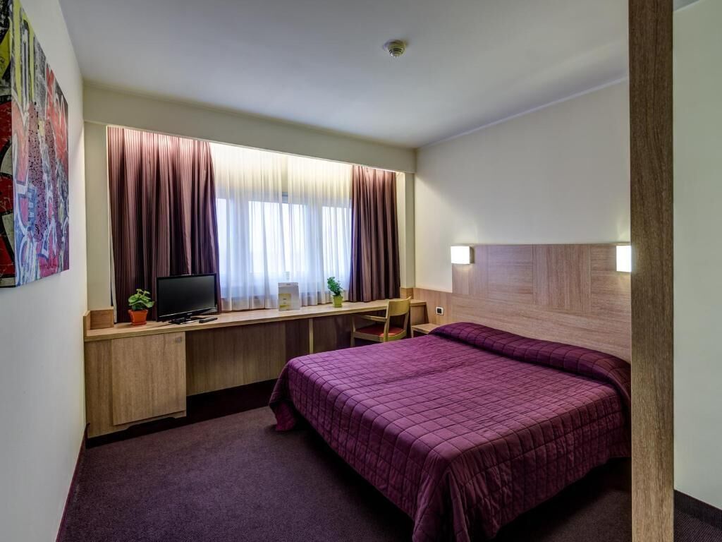 A hotel room with a bed, a desk with a computer, a chair, and a window with curtains.