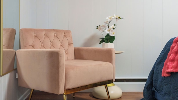 Una habitación con un sillón rosa, una mesa redonda blanca con una orquídea en maceta y una cama azul con una colcha rosa.
