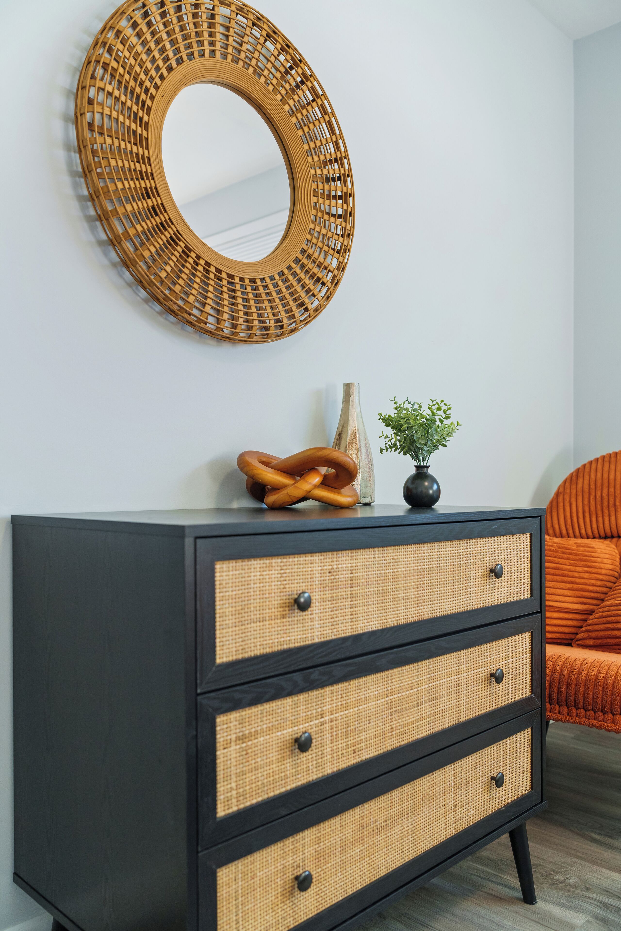 Une commode noire à trois tiroirs, un miroir à motifs tissés au-dessus, et une petite plante en pot posée sur le dessus.