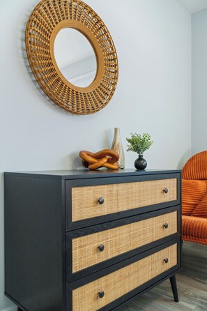 A black dresser with three drawers, a woven mirror above it, and a small potted plant on top.
