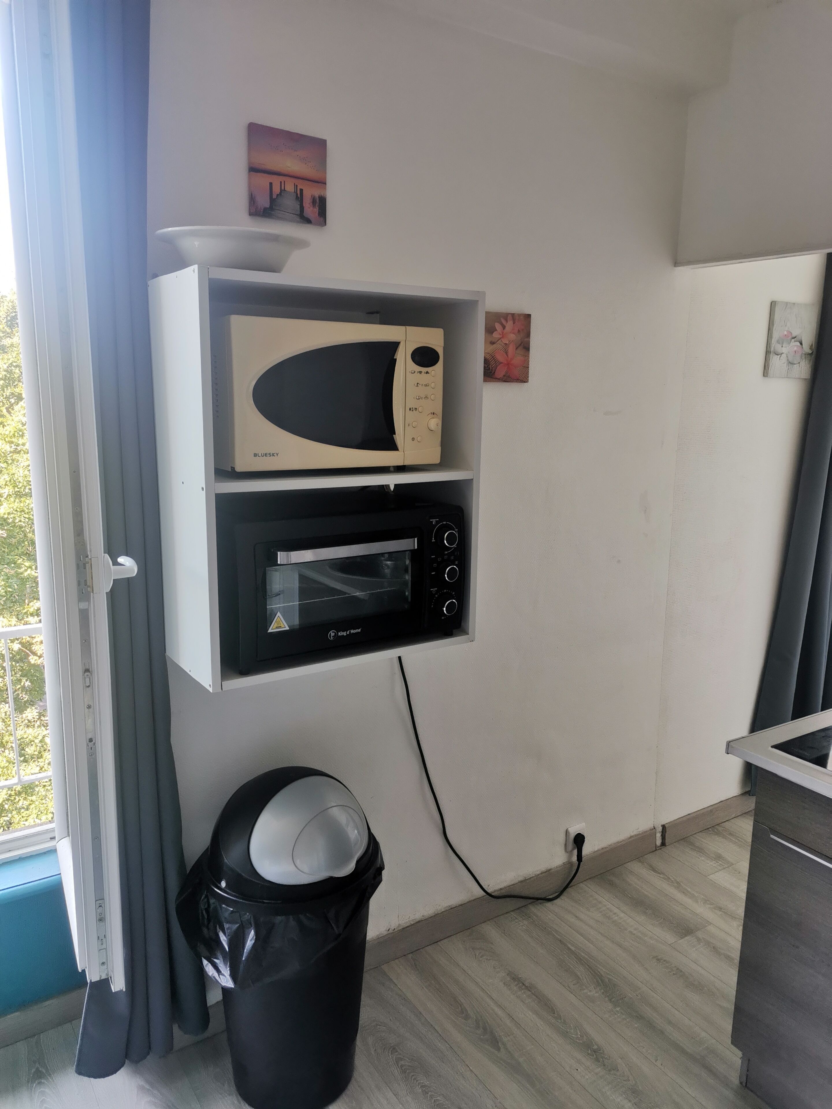 Una kitchenette con microondas y horno tostador fijados en la pared, un cubo de basura y una ventana con cortinas.
