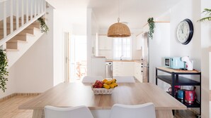 A modern kitchen with a wooden dining table, white chairs, a hanging basket with fruits, a clock, and a microwave. - Casa Da Roca Medano - Beach House & Terrace (Granadilla de Abona)
