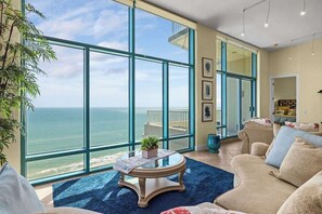 A spacious living room with large windows overlooking the ocean, a round coffee table with a plant, and a comfortable beige sofa. - Sapphire Penthouse Jr - Unrivaled Luxury - Views (South Padre Island)