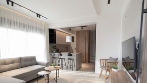Una sala moderna con un sofá, una mesa de centro y un televisor. Hay una zona de cocina con una barra y taburetes.