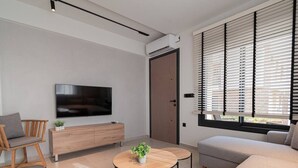 A modern living room with a flat-screen TV, a wooden cabinet, a round coffee table with a plant, a grey sofa, and a window with blinds. - Theano Sea Suite 3 by Travel Pro Services (Portaria)