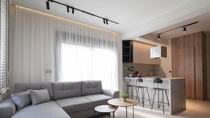A modern living room with a grey sectional sofa, a small round table, and a kitchen area with a bar counter and stools. - Theano Sea Suite 3 by Travel Pro Services (Portaria)