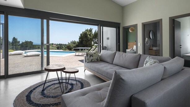A modern living room with a large sofa, a coffee table, and a view of a swimming pool and greenery outside.