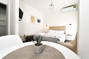 A modern hotel room with a bed, bedside table, and a small desk. There is a potted plant on the table, and a chair is visible in the foreground.