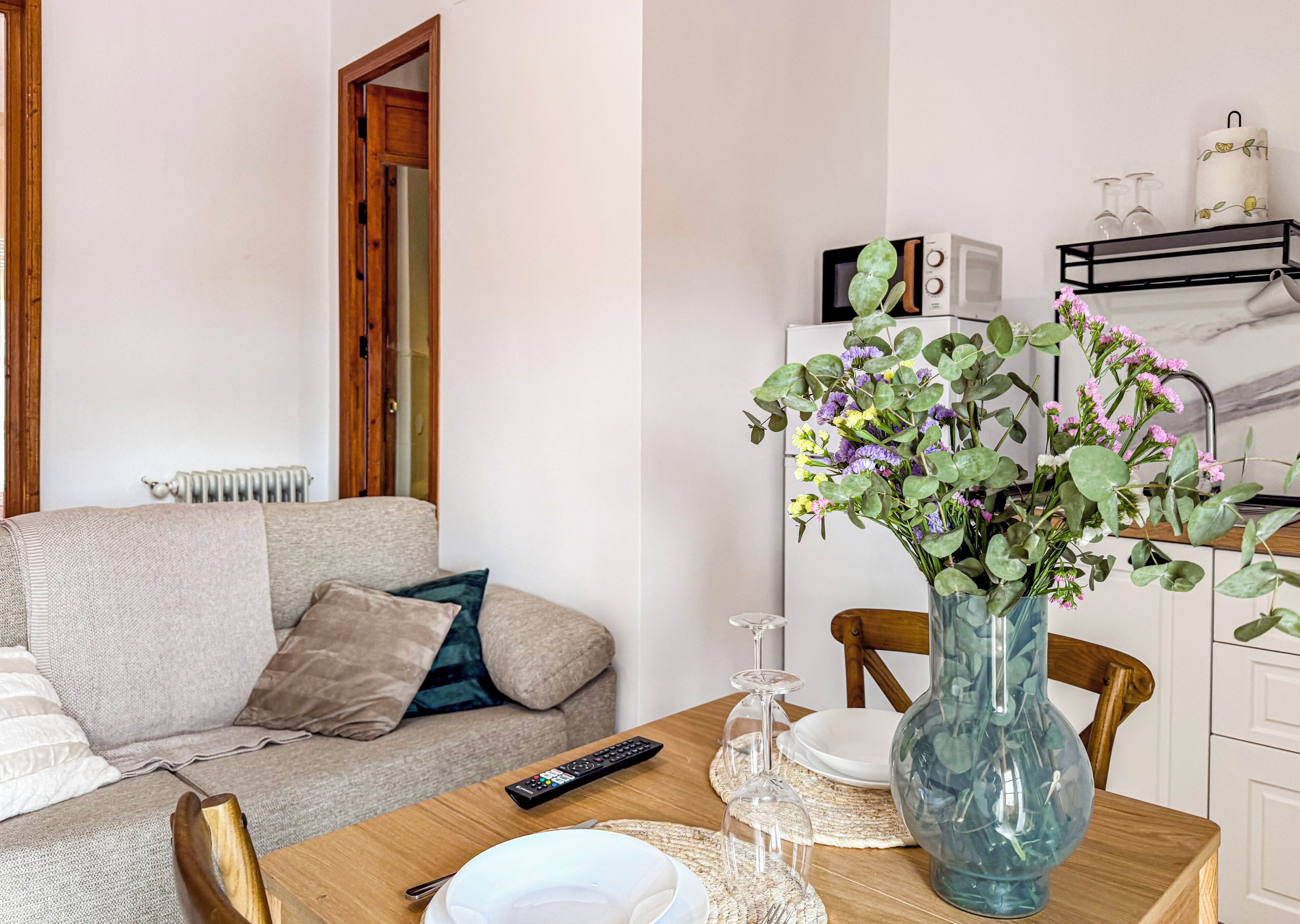 A living room with a sofa, a wooden dining table with a vase of flowers, a remote control, and a microwave on the counter.