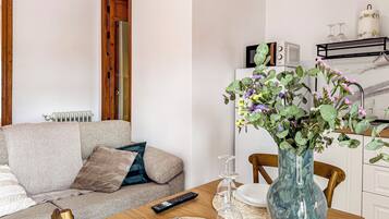 A living room with a sofa, a wooden dining table with a vase of flowers, a remote control, and a microwave on the counter.