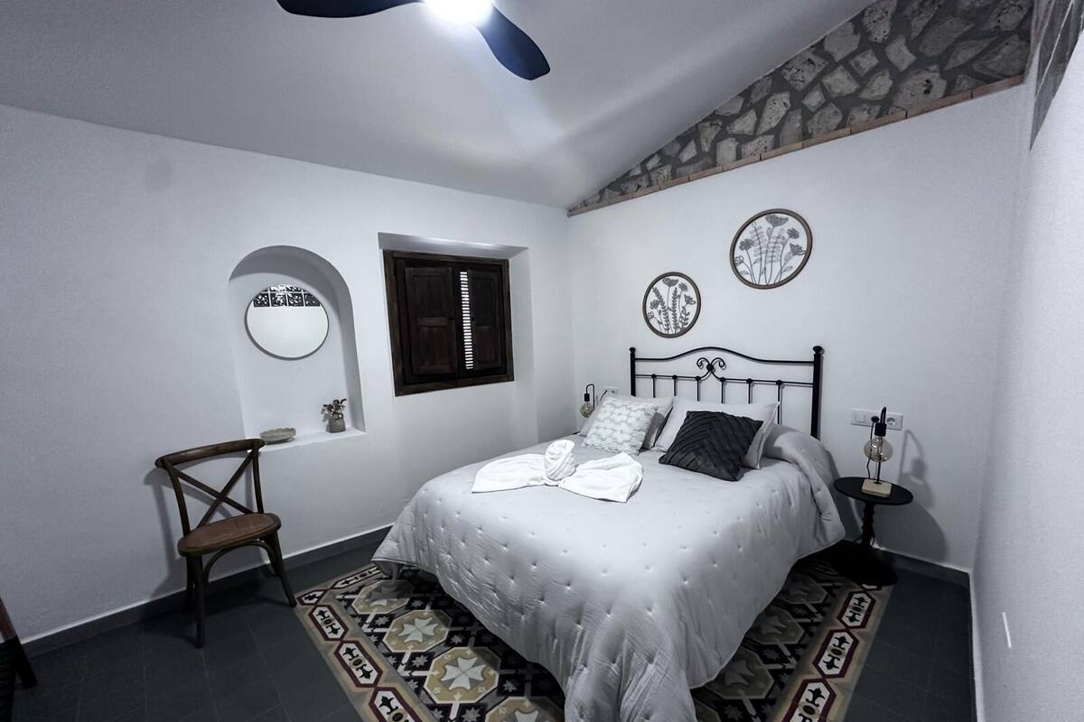 Une chambre à coucher comprenant un lit, une chaise, une petite table, un miroir fixé au mur et une fenêtre avec des volets.