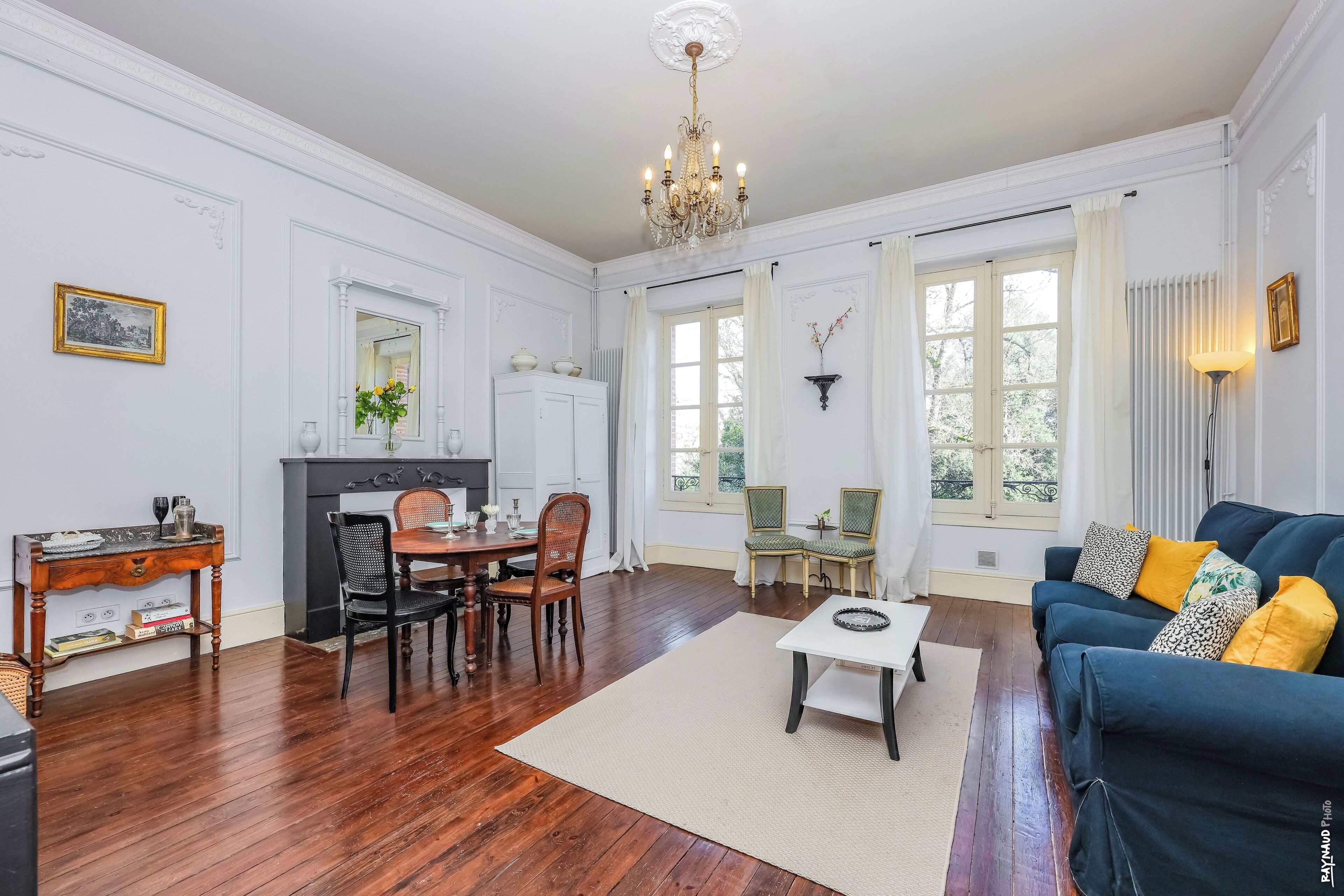 A room with a dining table, chairs, a sofa, a chandelier, and a fireplace.