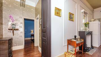 Un couloir doté d’un plancher en bois, d’une porte sombre, d’une lampe fixée au mur, d’une petite table avec une lampe et d’une armoire blanche agrémentée d’un miroir et d’objets décoratifs.