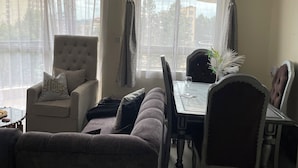 A living room with a sofa, armchair, dining table, and chairs. There is a window with curtains and a view of buildings outside. - Paradise Villa just outside Paradise/Karura forest for Nature visit,walks & more (Kiambu)