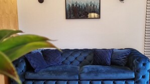 A blue tufted sofa with two cushions, a painting of a forest scene, and a potted plant in the foreground. - Comfortable guest house – 5 rooms wth private bathroom, Wi-Fi, kitchen & parking (Meine)