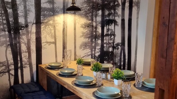 A dining area with a long wooden table set for four, surrounded by a forest-themed wall mural. - Comfortable guest house – 5 rooms wth private bathroom, Wi-Fi, kitchen & parking (Meine)