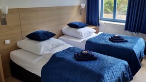 A hotel room with two beds, each with a blue quilted bedspread and pillows, a wooden headboard, a bedside lamp, and a view of greenery through the window. - Comfortable guest house – 5 rooms wth private bathroom, Wi-Fi, kitchen & parking (Meine)
