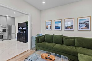 En moderne stue med en grøn sofa, et glasbord og et køkken i baggrunden.