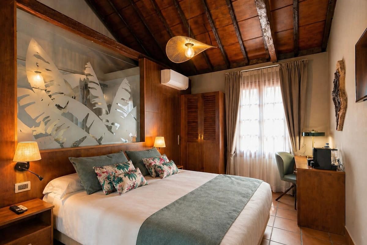 Une chambre à coucher comprenant un lit, des tables de chevet, un bureau, une chaise, une grande fenêtre avec des rideaux et un ventilateur de plafond.