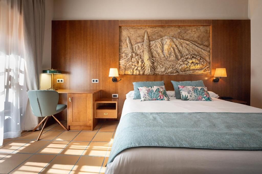 A hotel room with a bed, desk, chair, lamps, and a large wall painting.