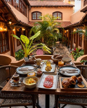 A dining table set with various dishes, surrounded by wicker chairs, in a courtyard with plants and a building in the background.. Hotel Jardin Secreto
