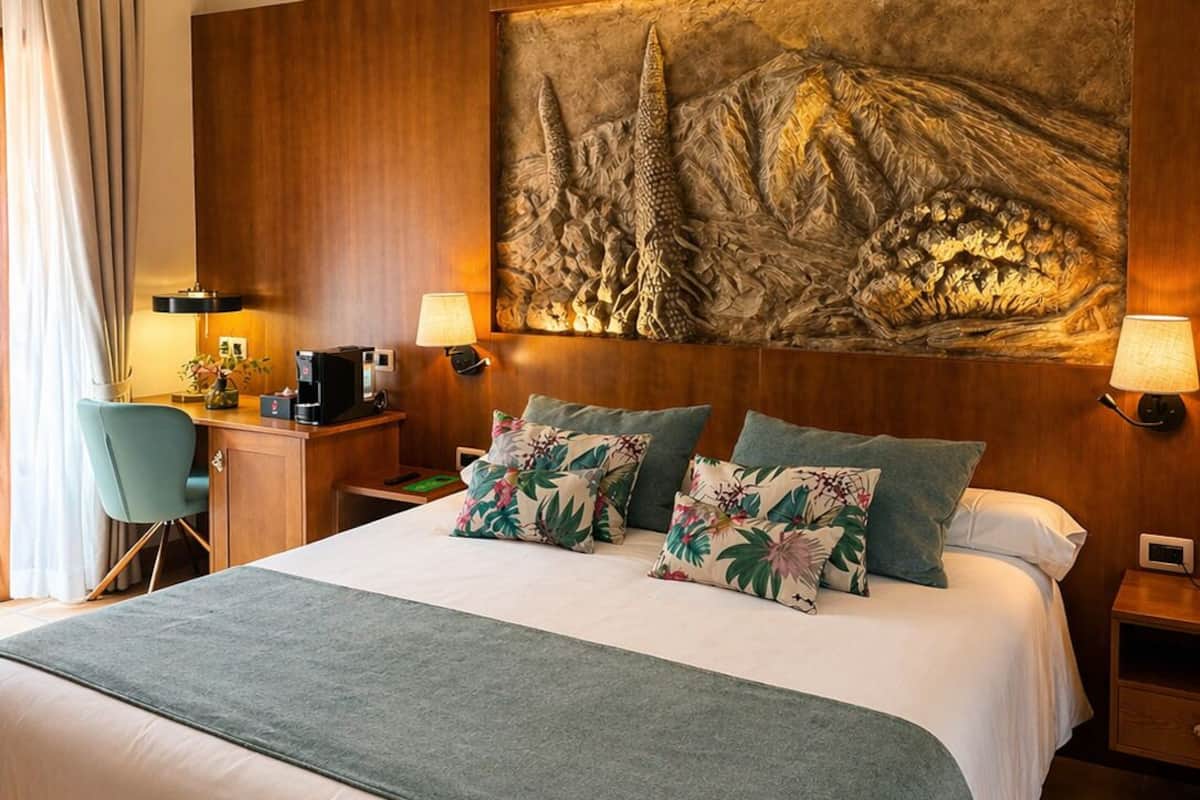 Une chambre d’hôtel avec un grand lit, deux tables de chevet, un bureau avec une chaise, une grande fresque murale et une vue sur l’extérieur.