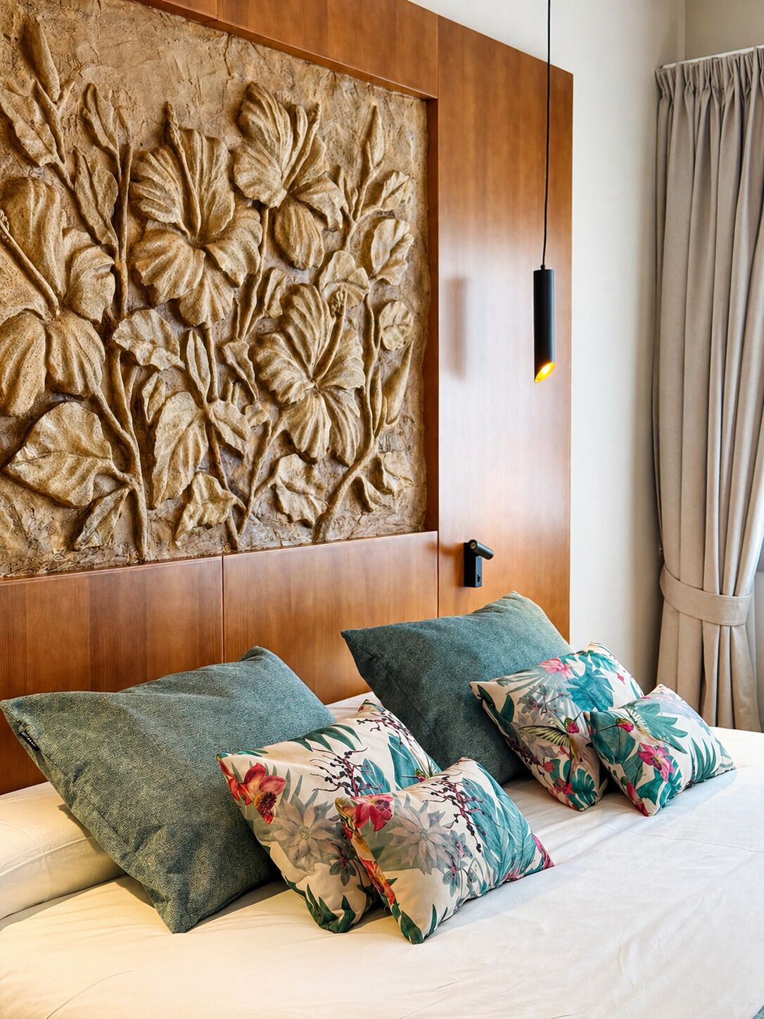 Habitación de hotel con cabecera de madera, almohadas estampadas con motivos florales y una lámpara fijada a la pared.