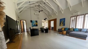 A spacious living area with a large TV, a dining table, and a sofa. There are ceiling fans and artwork on the walls.