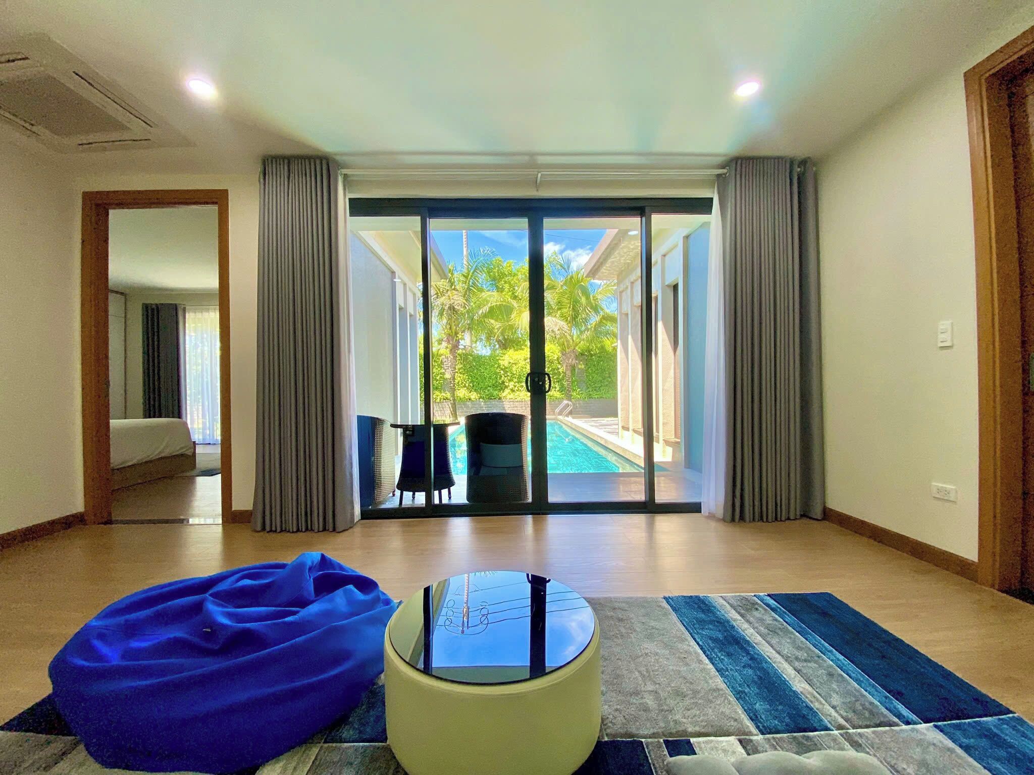 Una habitación de hotel con un amplio ventanal corredero que da a una zona de piscina, un sillón orejero azul y una pequeña mesa redonda.