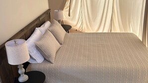 En seng med et vamset tæppe, en sengetavle og et lille rundt bord.