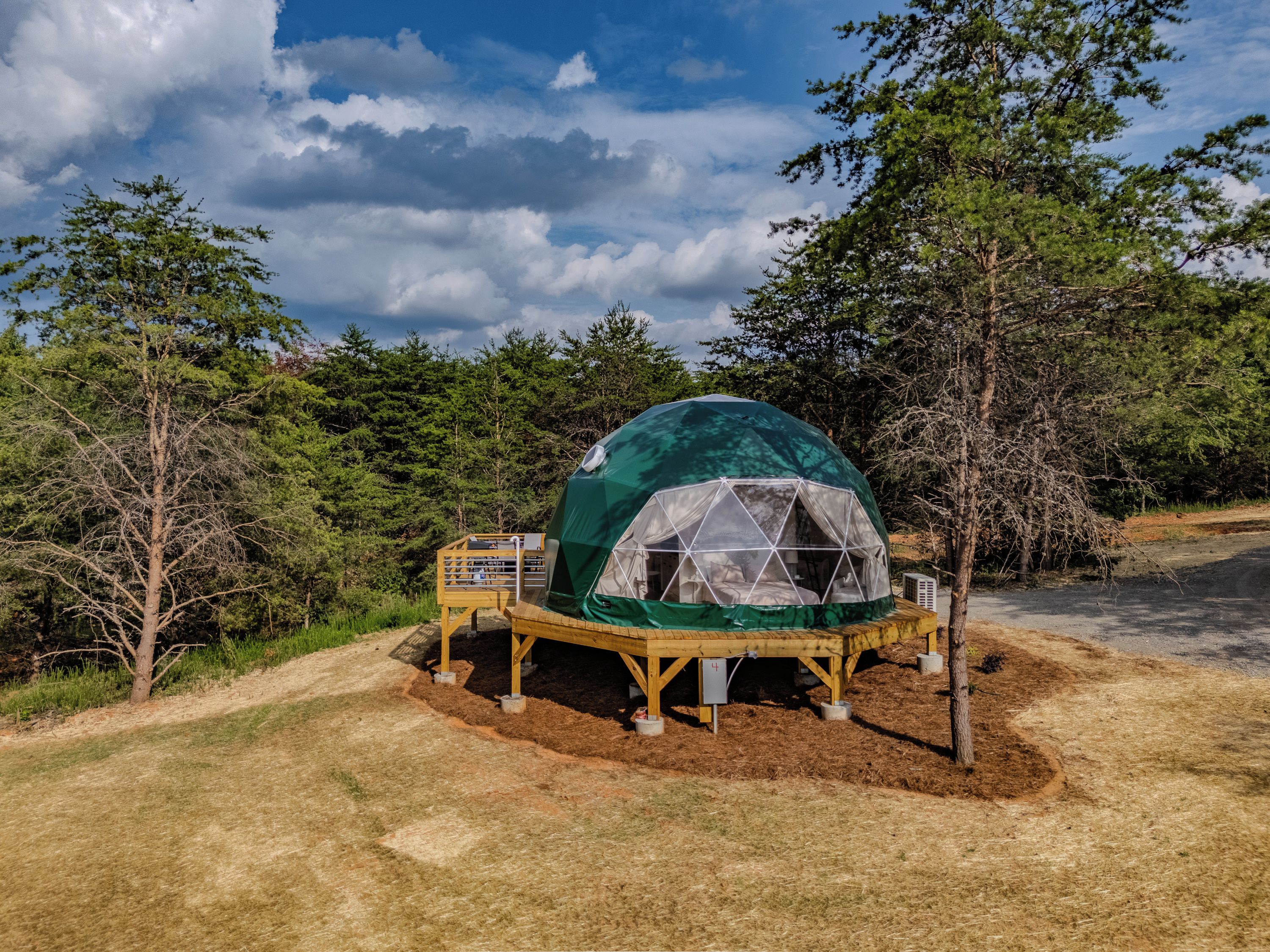 Een geodetische koepeltent met een houten terras, omgeven door bomen en een heldere lucht.
