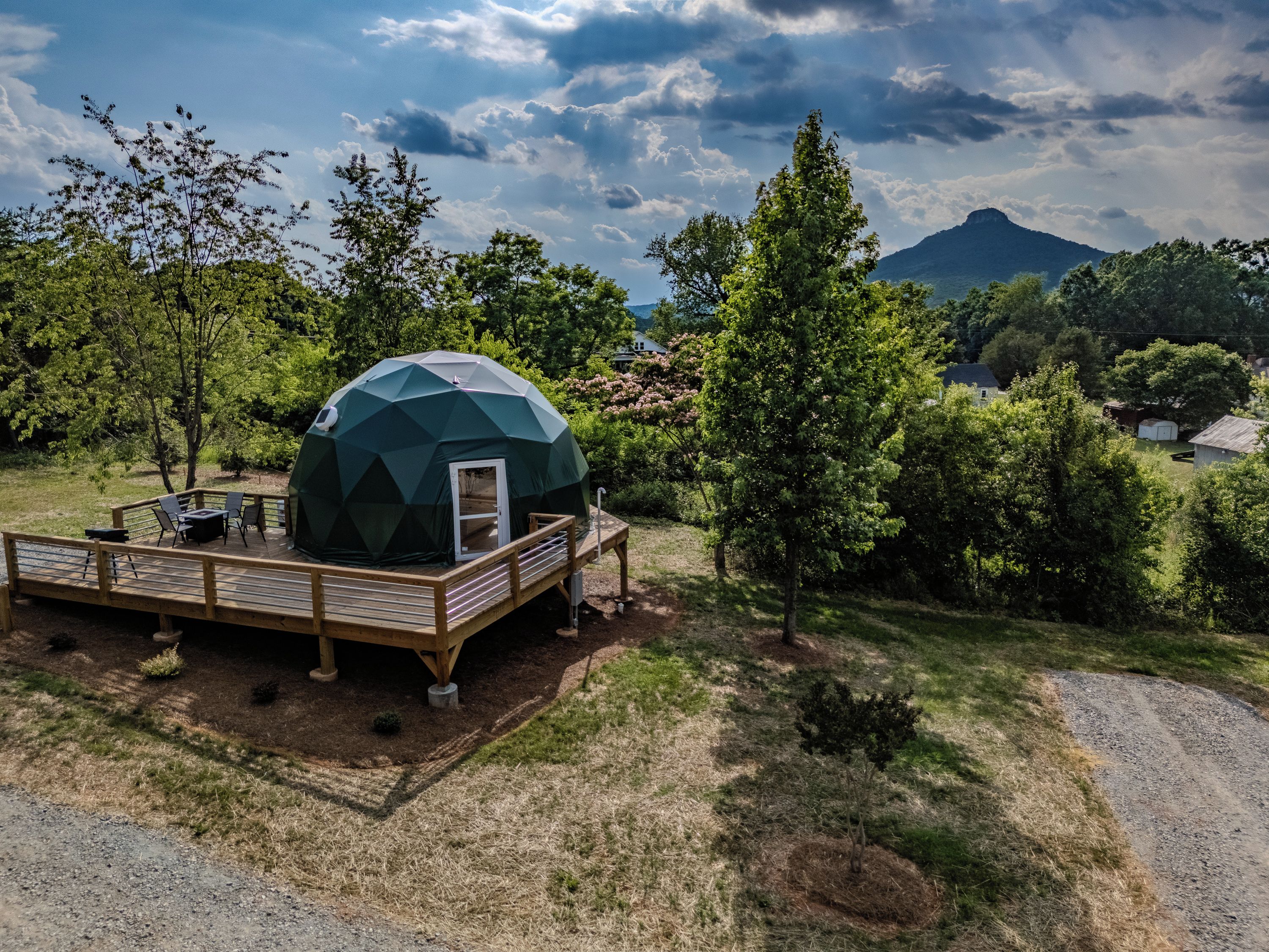 A geodesic dome structure with a deck and outdoor seating, surrounded by trees and a mountain in the background.