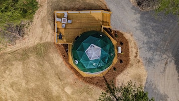 Eine Vogelperspektive zeigt eine Holzterrasse mit einer kreisförmigen, grünen Zeltdachkonstruktion, umgeben von einem unbefestigten Weg und spärlicher Vegetation.